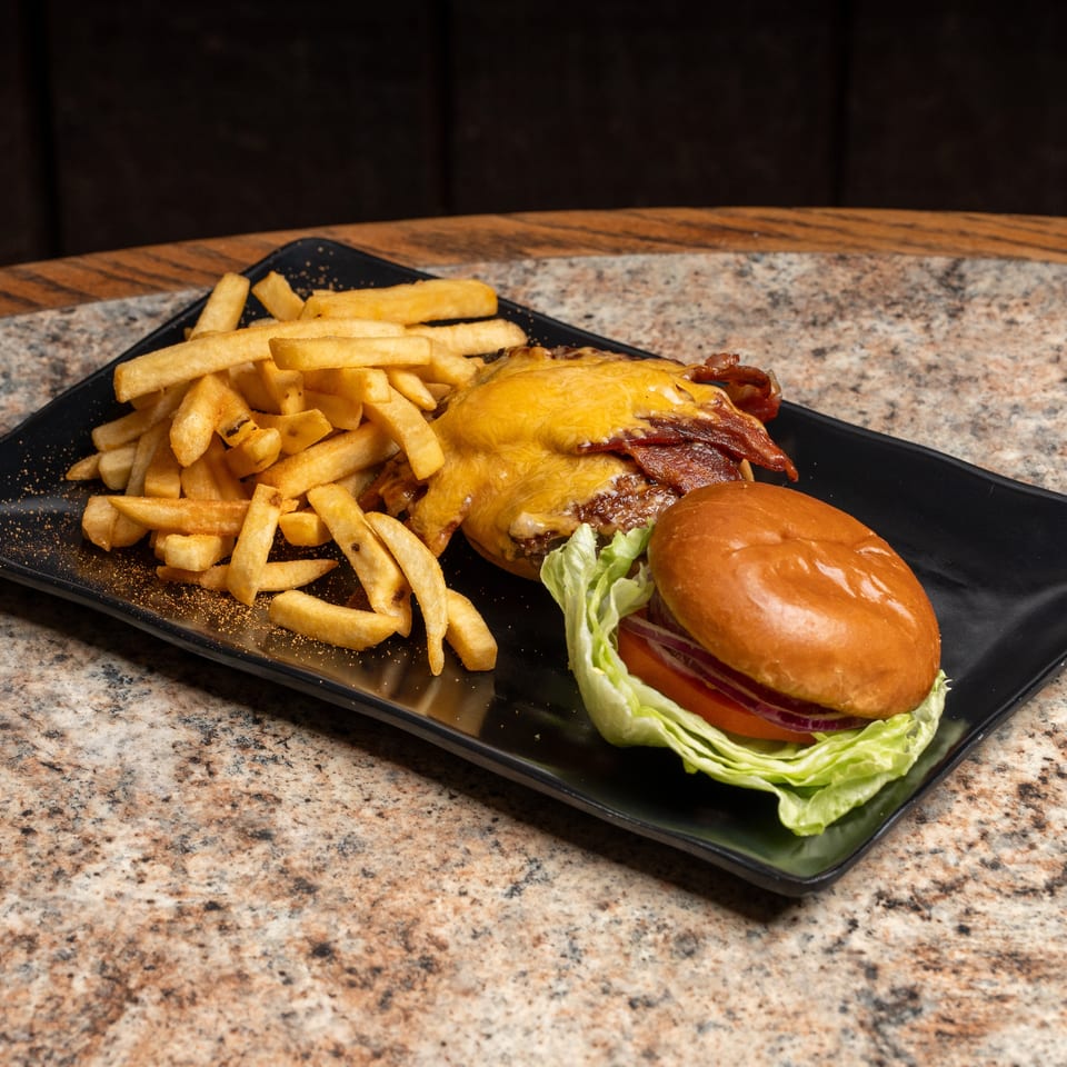 Cheesy bacon burger with lettuce, tomato, and red onion, served with seasoned fries on a black plate.