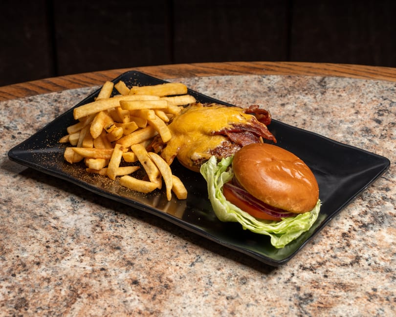 Cheesy bacon burger with lettuce, tomato, and red onion, served with seasoned fries on a black plate.