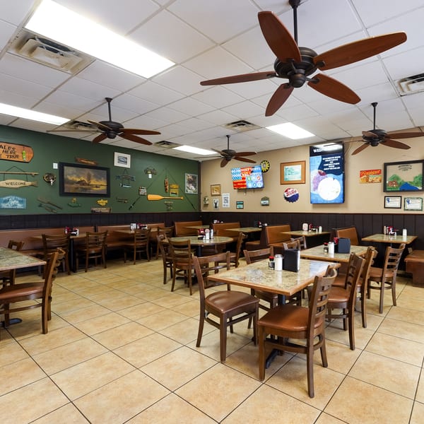 Restaurant dining room with fishing-themed decor, ceiling fans, wooden tables, chairs, and brown booth seating.