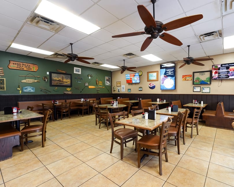 Restaurant dining room with fishing-themed decor, ceiling fans, wooden tables, chairs, and brown booth seating.