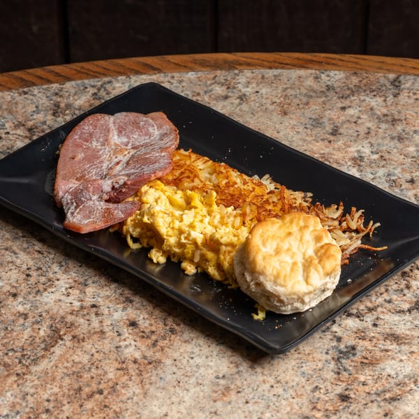 Breakfast plate featuring ham, scrambled eggs, crispy hash browns, and a fluffy biscuit.