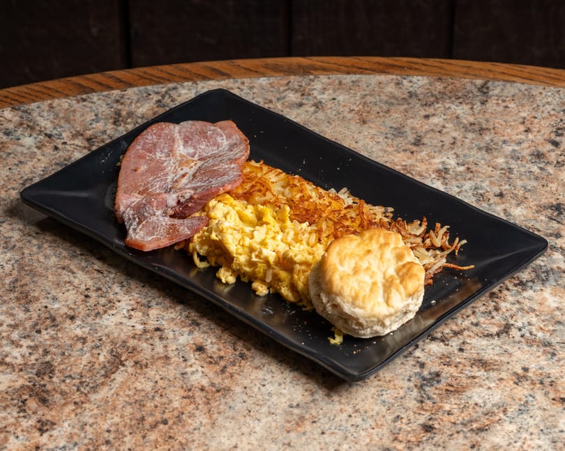Breakfast plate featuring ham, scrambled eggs, crispy hash browns, and a fluffy biscuit.