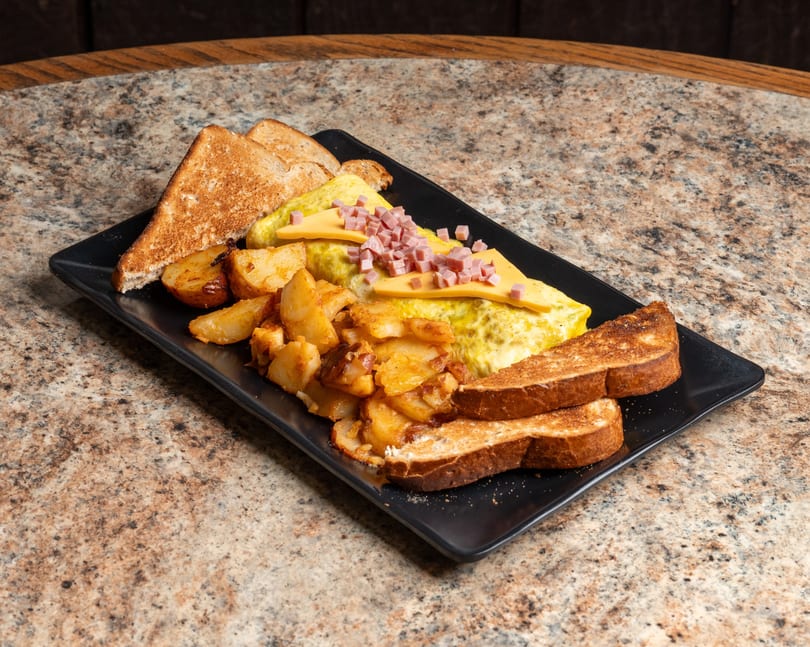 Hearty breakfast plate featuring a ham and cheese omelet, crispy potatoes, and golden toast.