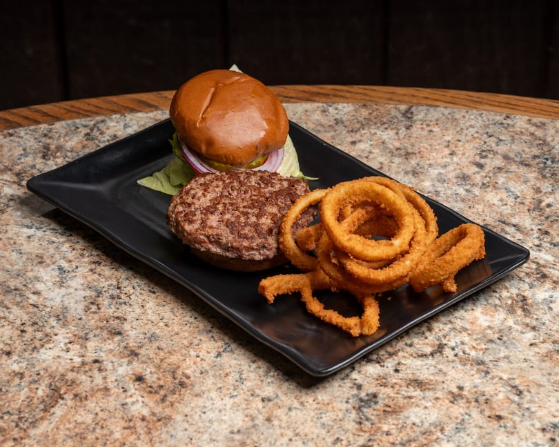 Juicy burger with lettuce, red onion, and pickle, alongside golden onion rings on a black plate.