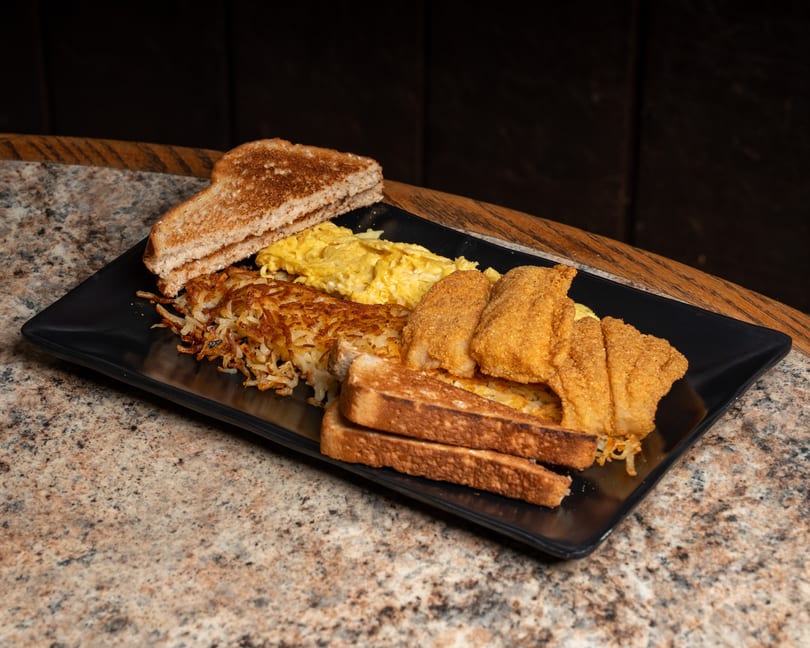 Breakfast plate with fried fish, scrambled eggs, hash browns, and toast on a black plate.