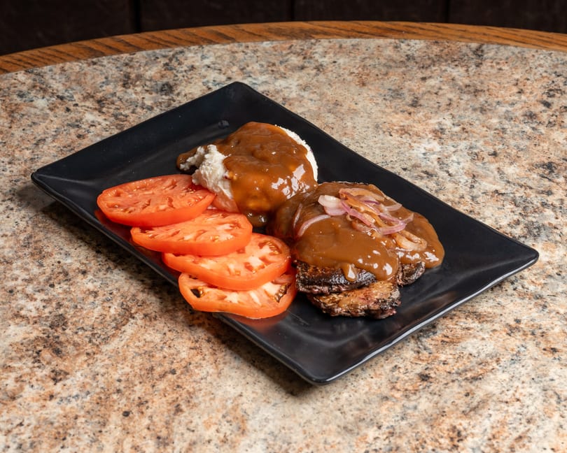 Grilled steak with gravy and onions, mashed potatoes, and sliced tomatoes on a black plate.