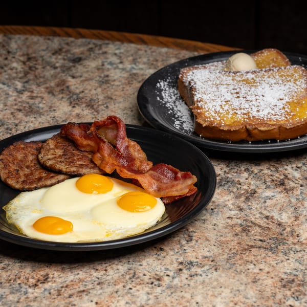 Breakfast plates with fried eggs, sausage, bacon, and powdered French toast with butter.