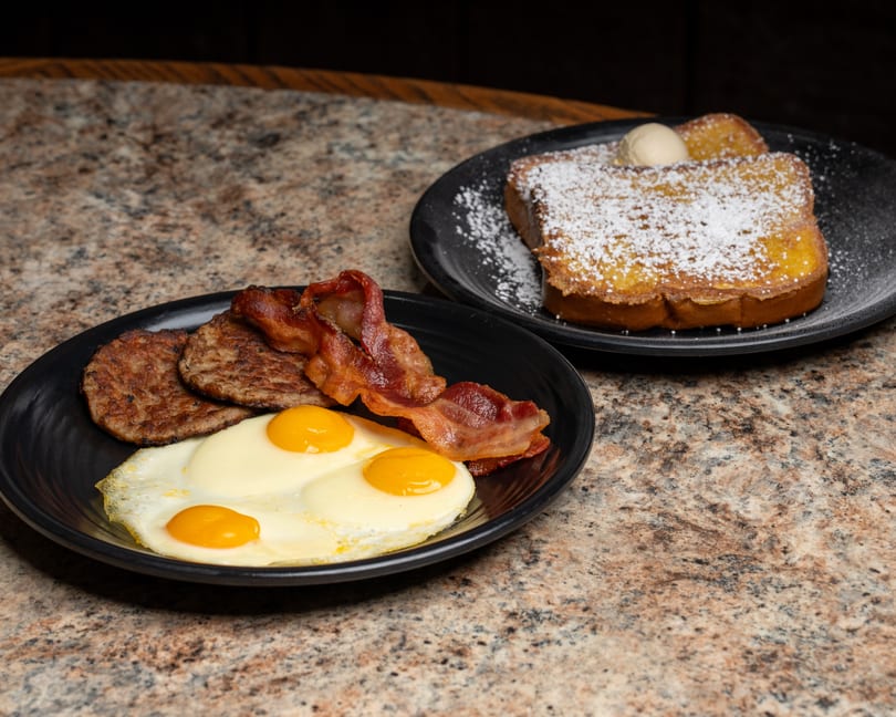 Breakfast plates with fried eggs, sausage, bacon, and powdered French toast with butter.