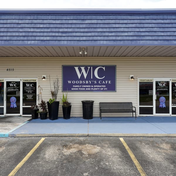 Woodsby's Cafe storefront with a blue roof, beige siding, entrance doors, outdoor bench, and planters.