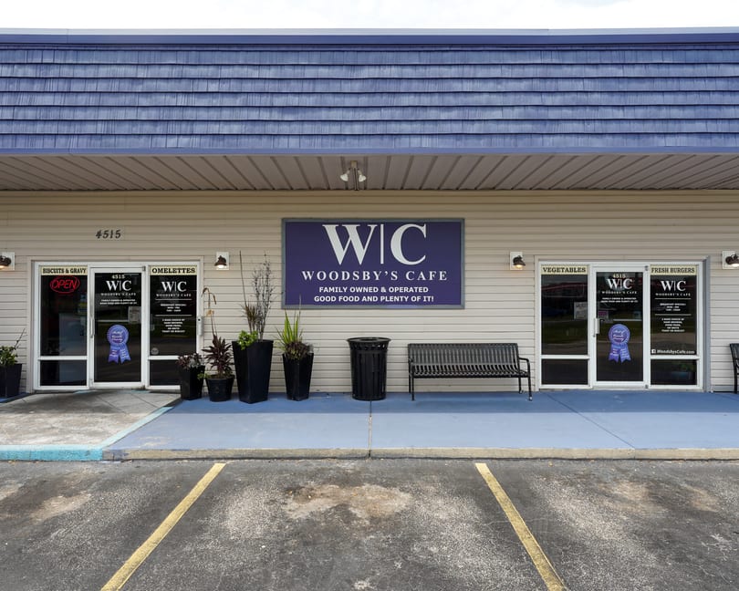 Woodsby's Cafe storefront with a blue roof, beige siding, entrance doors, outdoor bench, and planters.